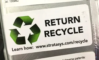 En etikett som sitter monterad på en spole filament. Det står "RETURN RECYCLE", en hänvisning att man kan återvinna spolarna till Stratasys.